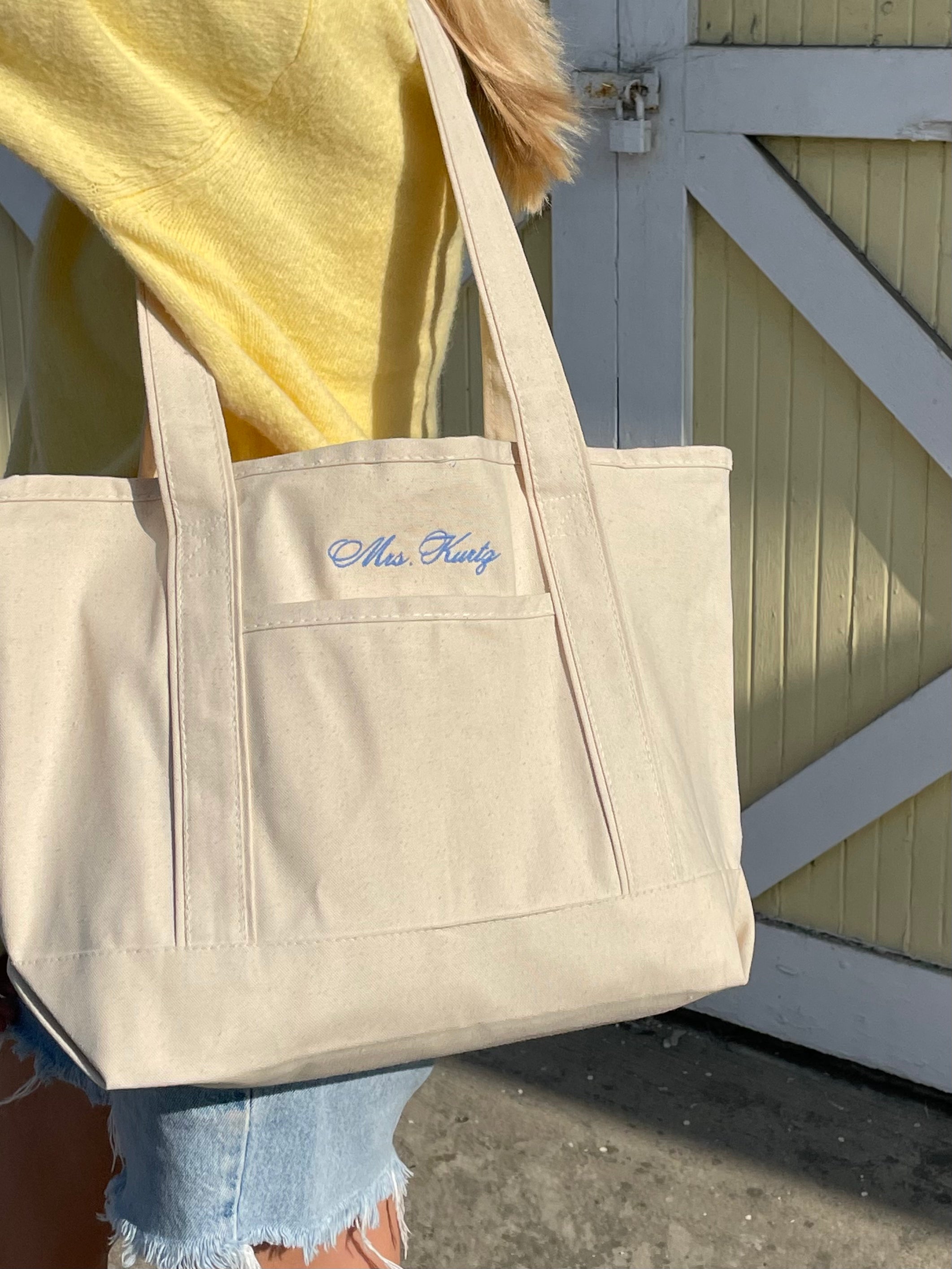 Beige tote bag with 'Mrs. Kurtz' embroidery held by a person wearing a yellow top.