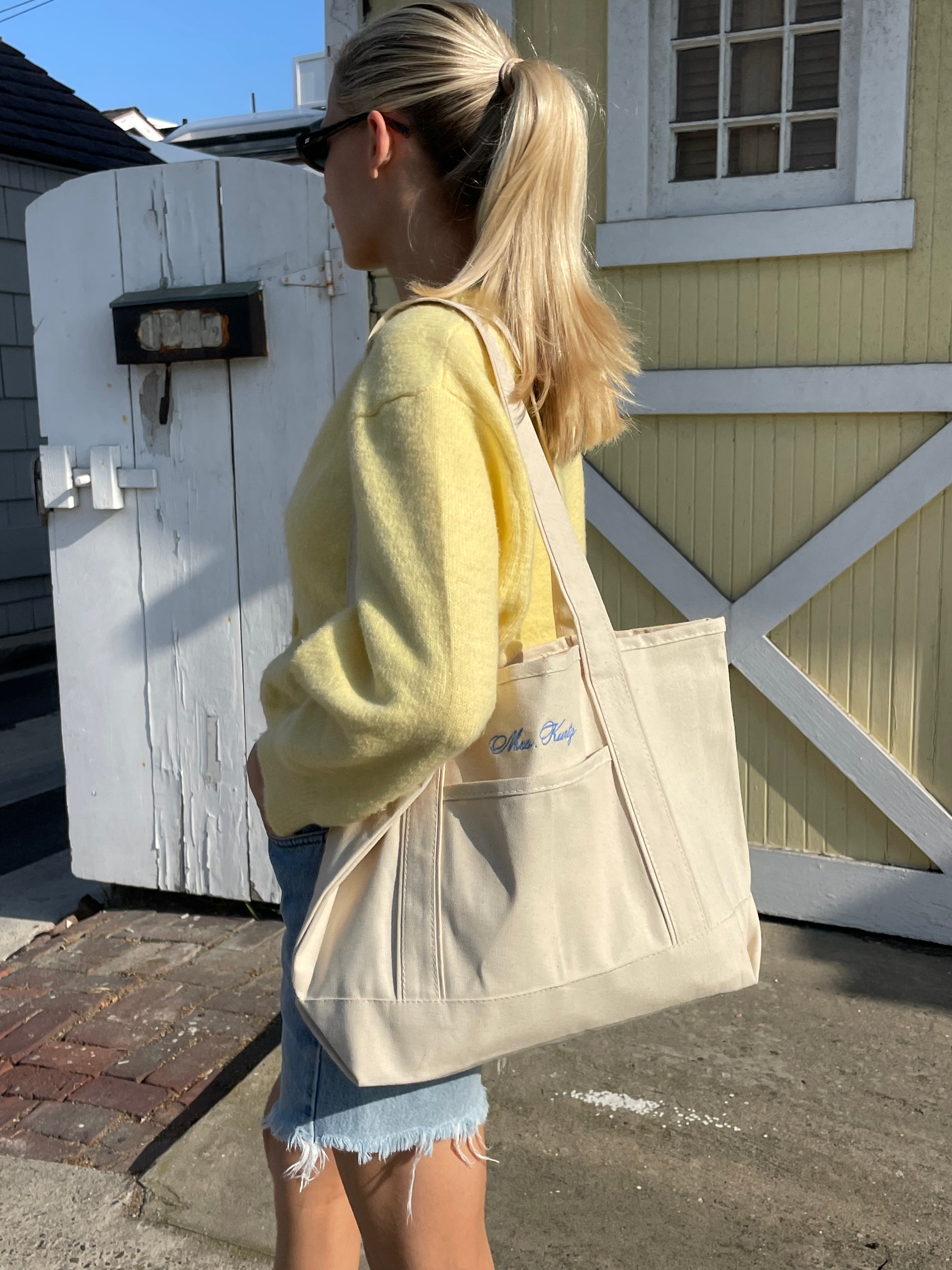 Person holding a beige tote bag with a brand logo outdoors.